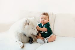 Cute baby in green shirt sitting indoors with a fluffy white Samoyed dog.