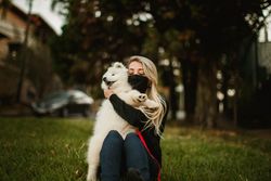 Relaxed female in medical mask sitting on lawn and cuddling adorable fluffy dog during stroll in park