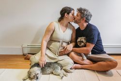 Full body of happy pregnant woman and man cuddling and kissing each other while resting on floor at home with cute Yorks
