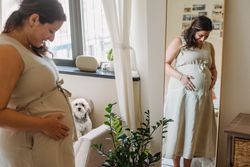 Side view of crop happy young pregnant female in casual dress smiling while touching belly standing in front of mirror n