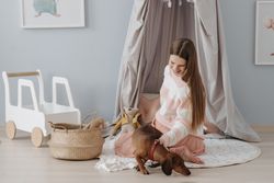 A smiling pregnant woman sits in a nursery, petting her dachshund dog.